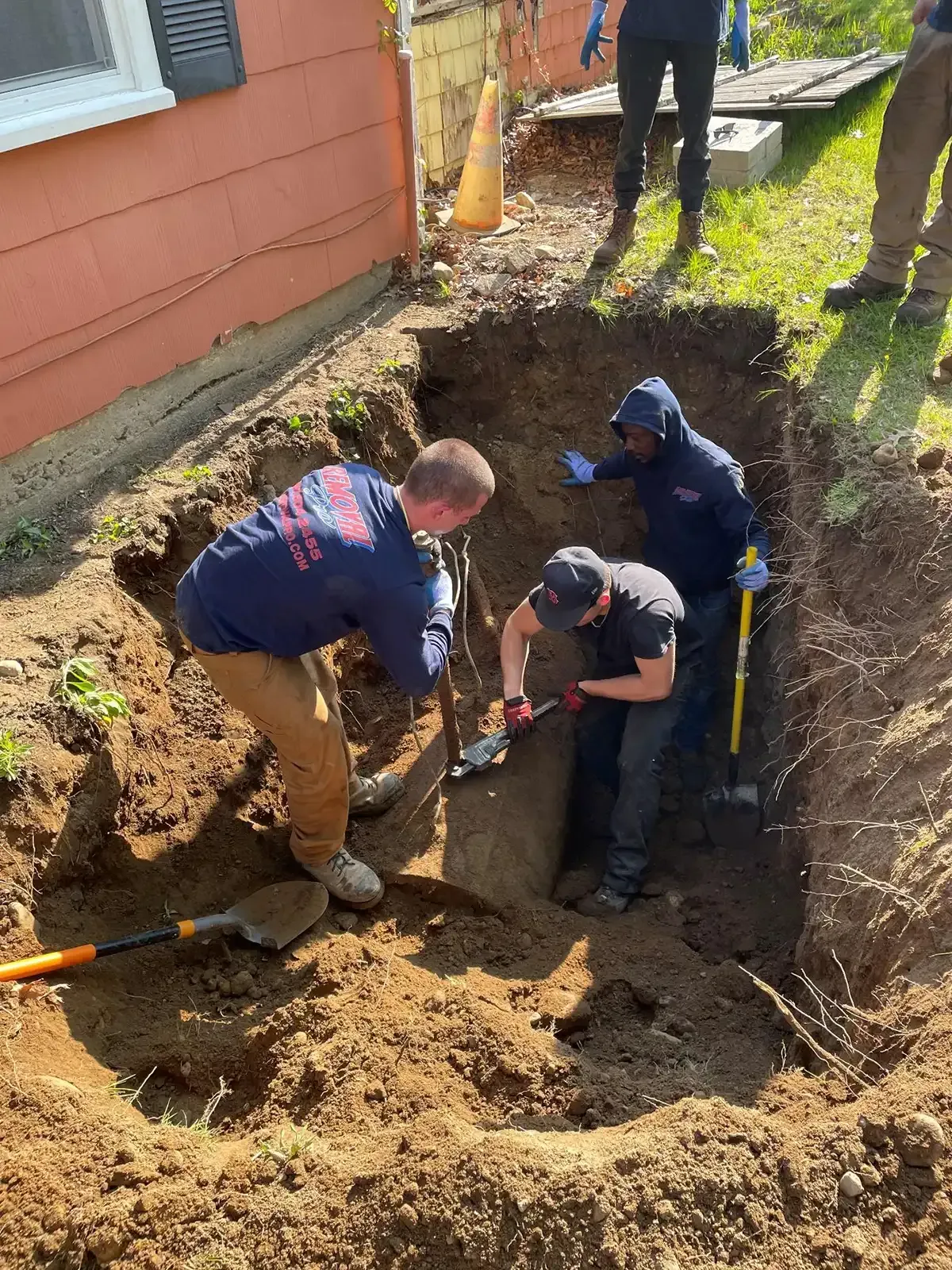 Workers repairing a trench beside a house, using tools in a dirt excavation site.
