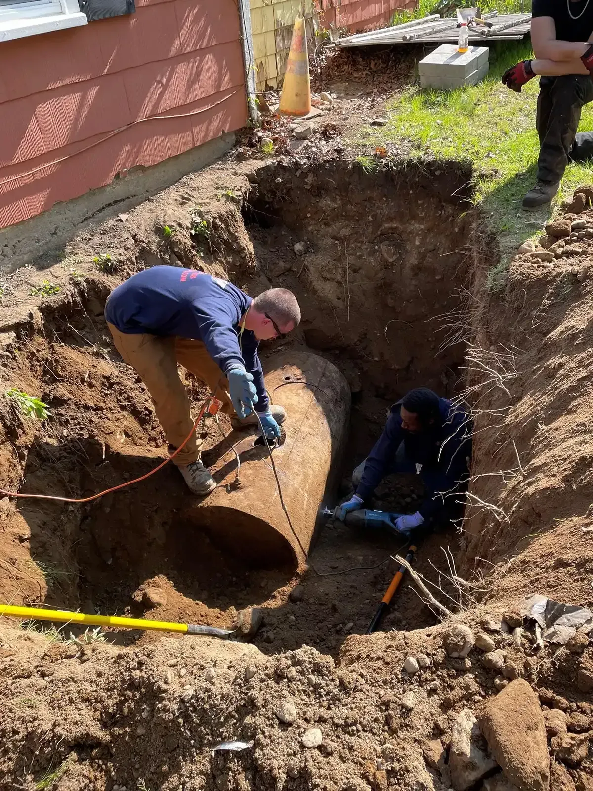Workers in a trench lifting a large buried tank near a house.