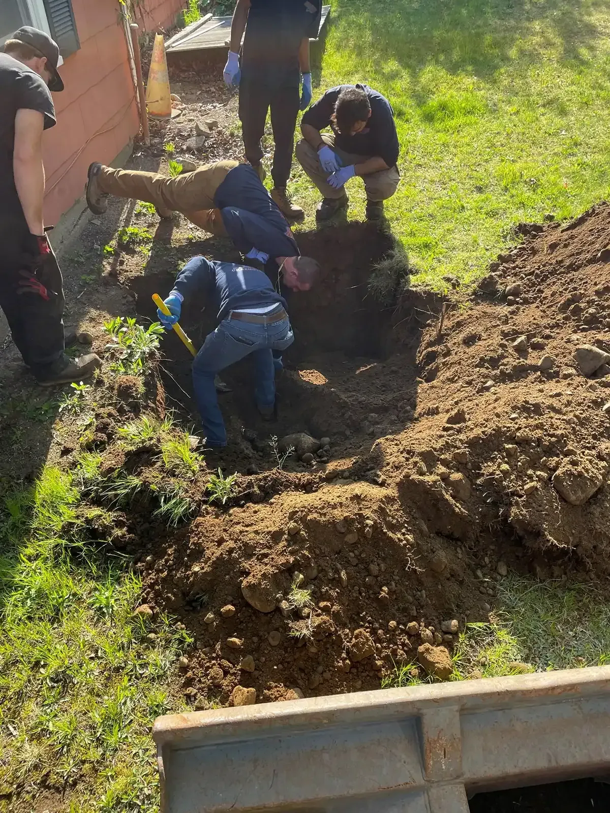 Workers digging a large hole beside a dirt trench near a grassy area.