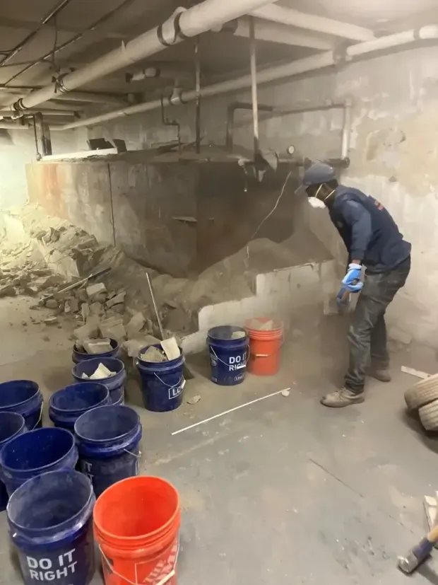 A worker in a mask and vest cleans around a large metal tank in a basement, using blue and orange buckets for debris.