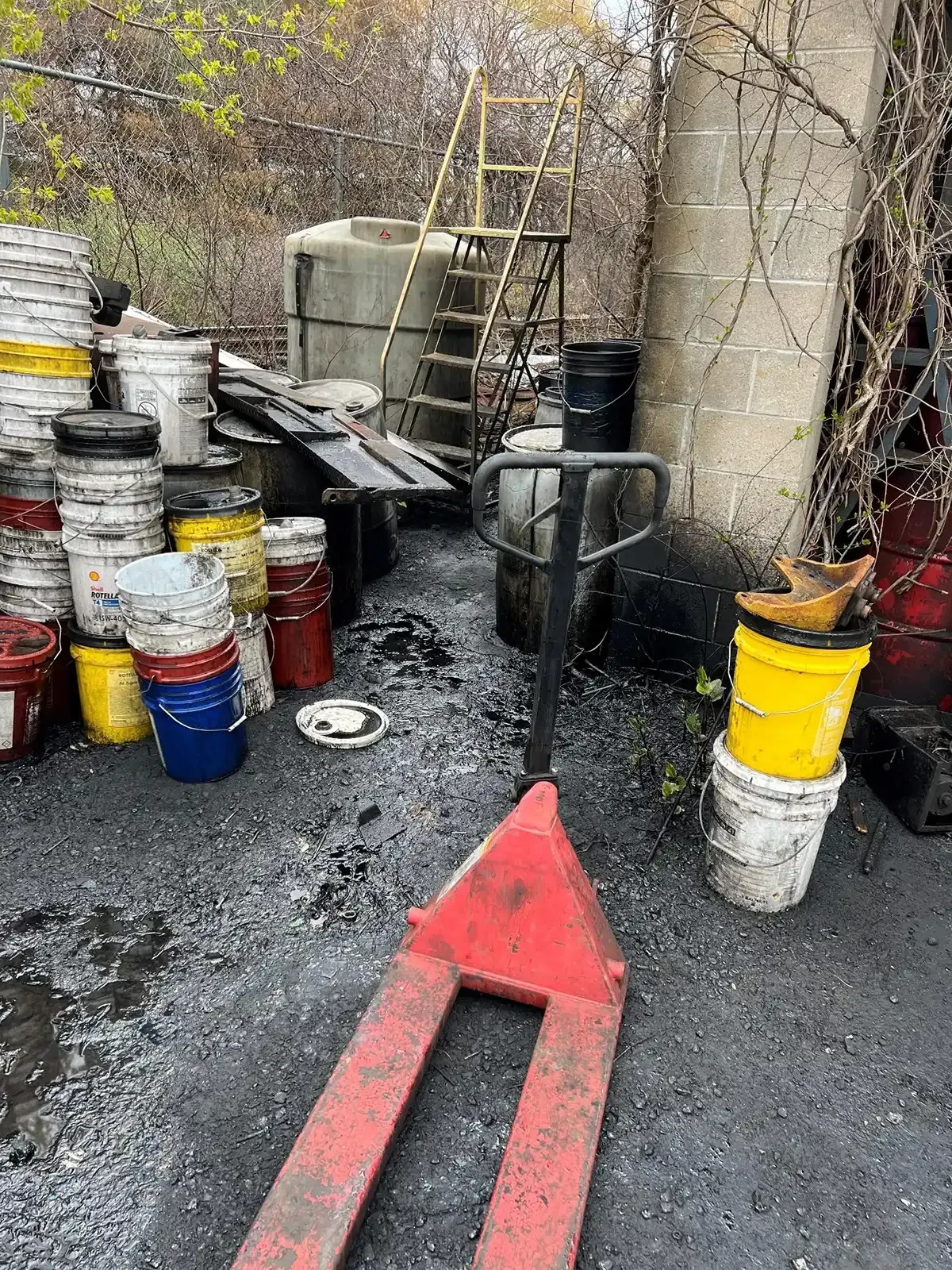 Outdoor work area with stacked paint buckets, a ladder, and a red pallet jack on wet gravel.