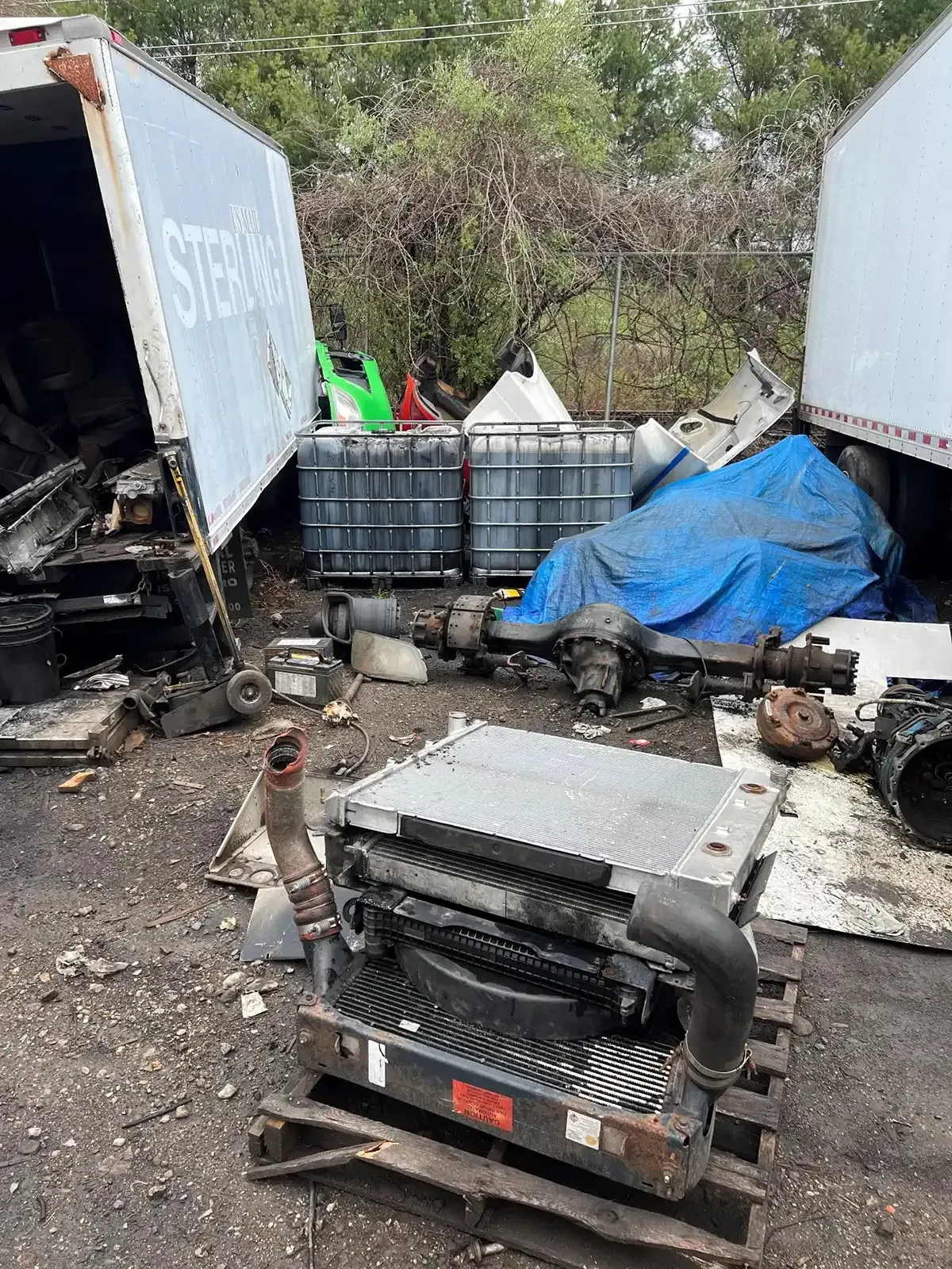 A cluttered scrapyard with damaged trucks, metal debris, and a blue tarp over stacked bins.