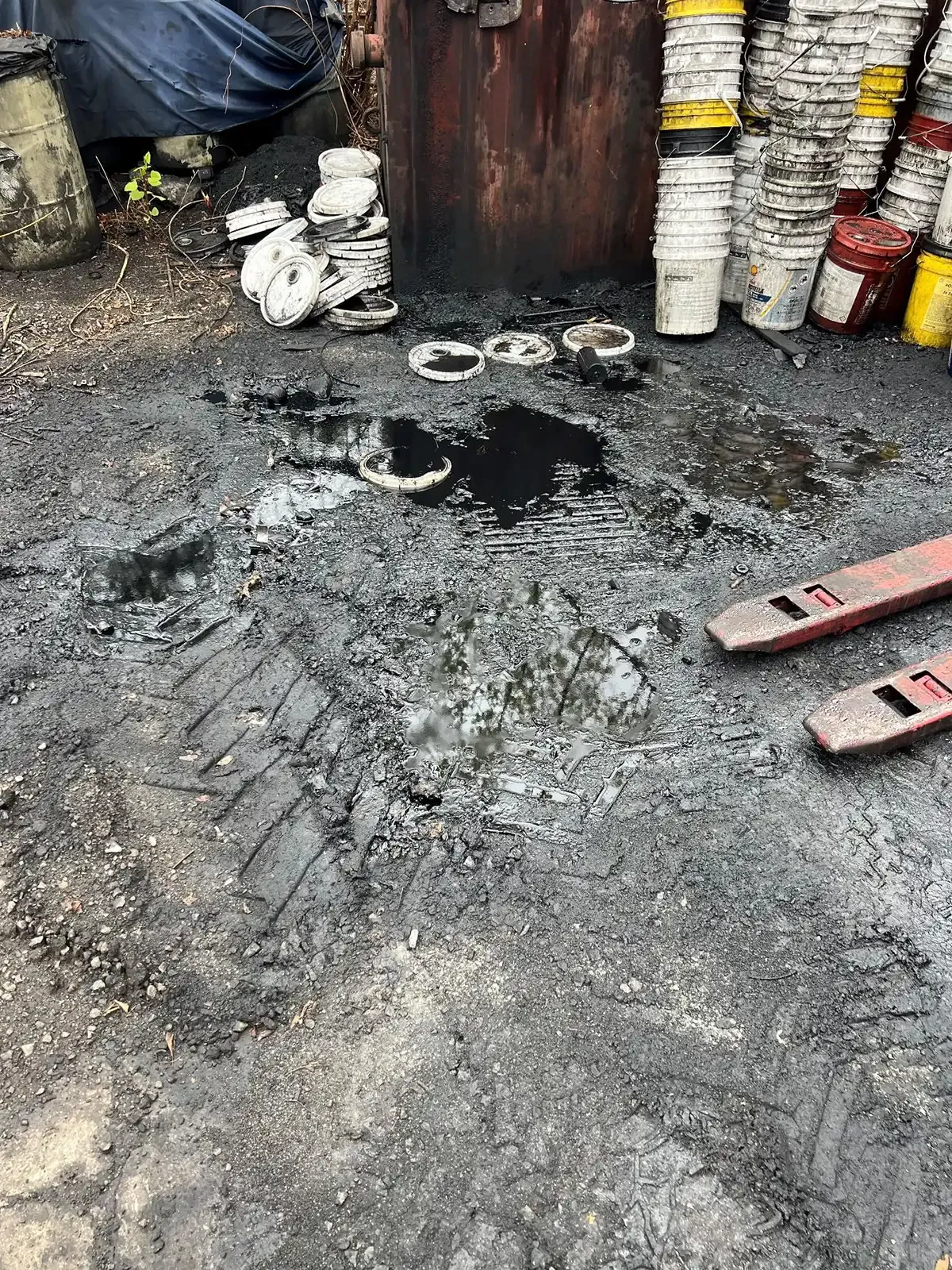 Charred, debris-strewn ground with black soot, buckets, and a red hand pallet jack near a metal door.