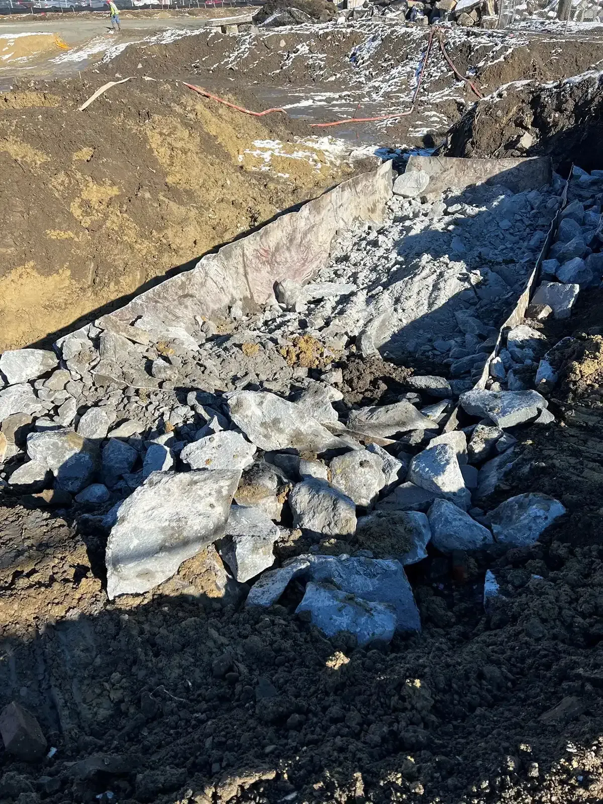 Pile of broken concrete and rubble beside a road embankment, with dirt and snow in bright sunlight.