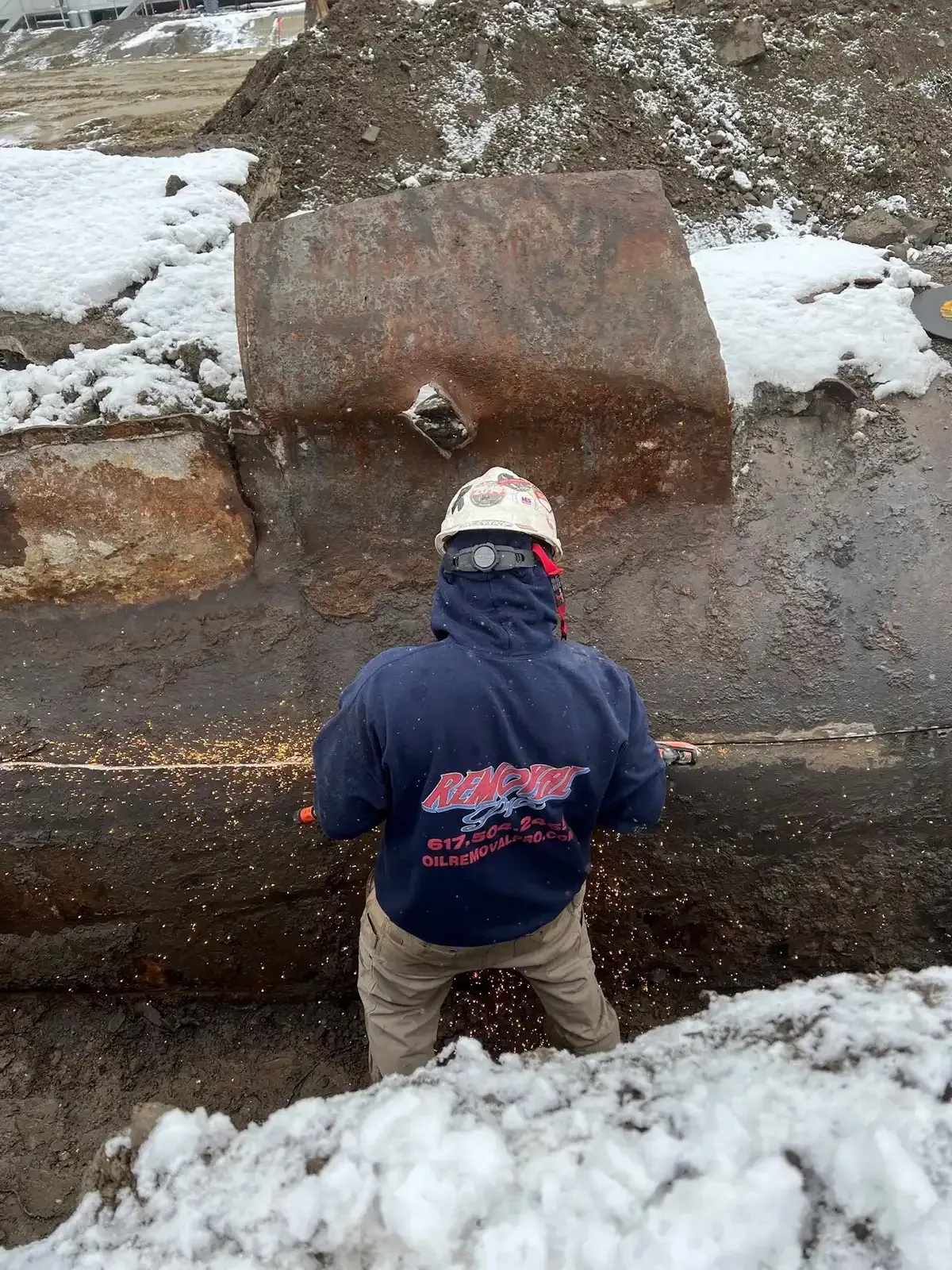 Worker in a hard hat stands by a large rusty metal pipe in snow.