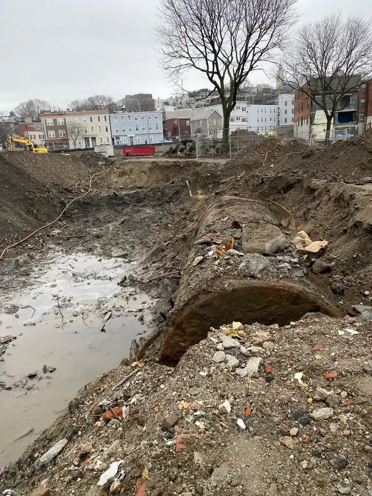 Large muddy excavation pit with standing water and debris, bare trees, and buildings in the background.