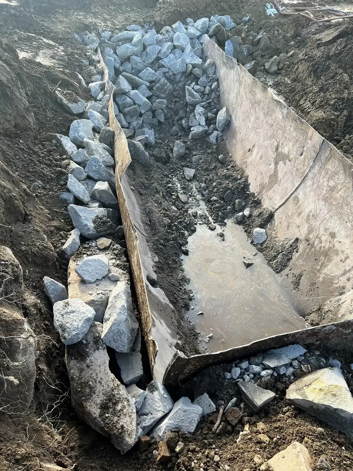 Rock-lined concrete drainage ditch with shallow muddy water on a dirt slope.