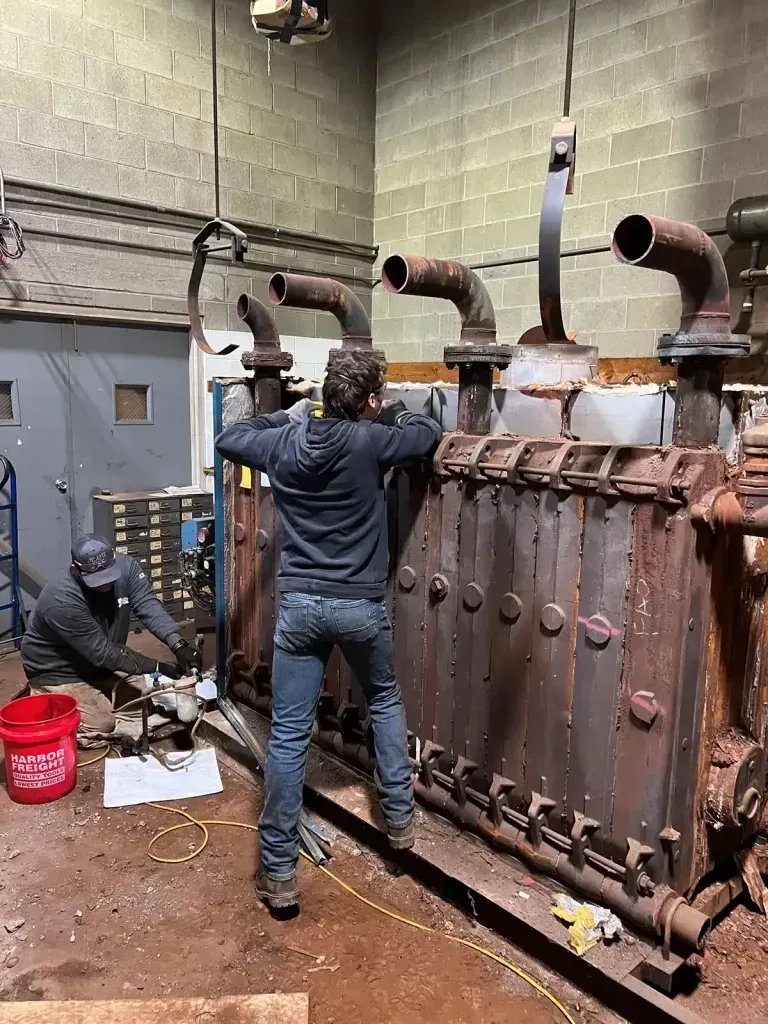 Two people work on a large, rusty industrial machine with four vertical pipes in a brick-walled workshop.