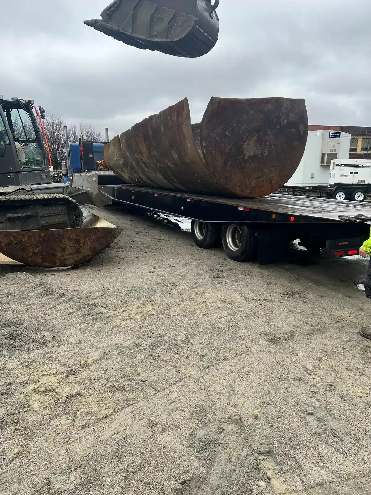 Excavator loading a large rusted metal bucket onto a flatbed trailer in a gravel yard under cloudy skies.