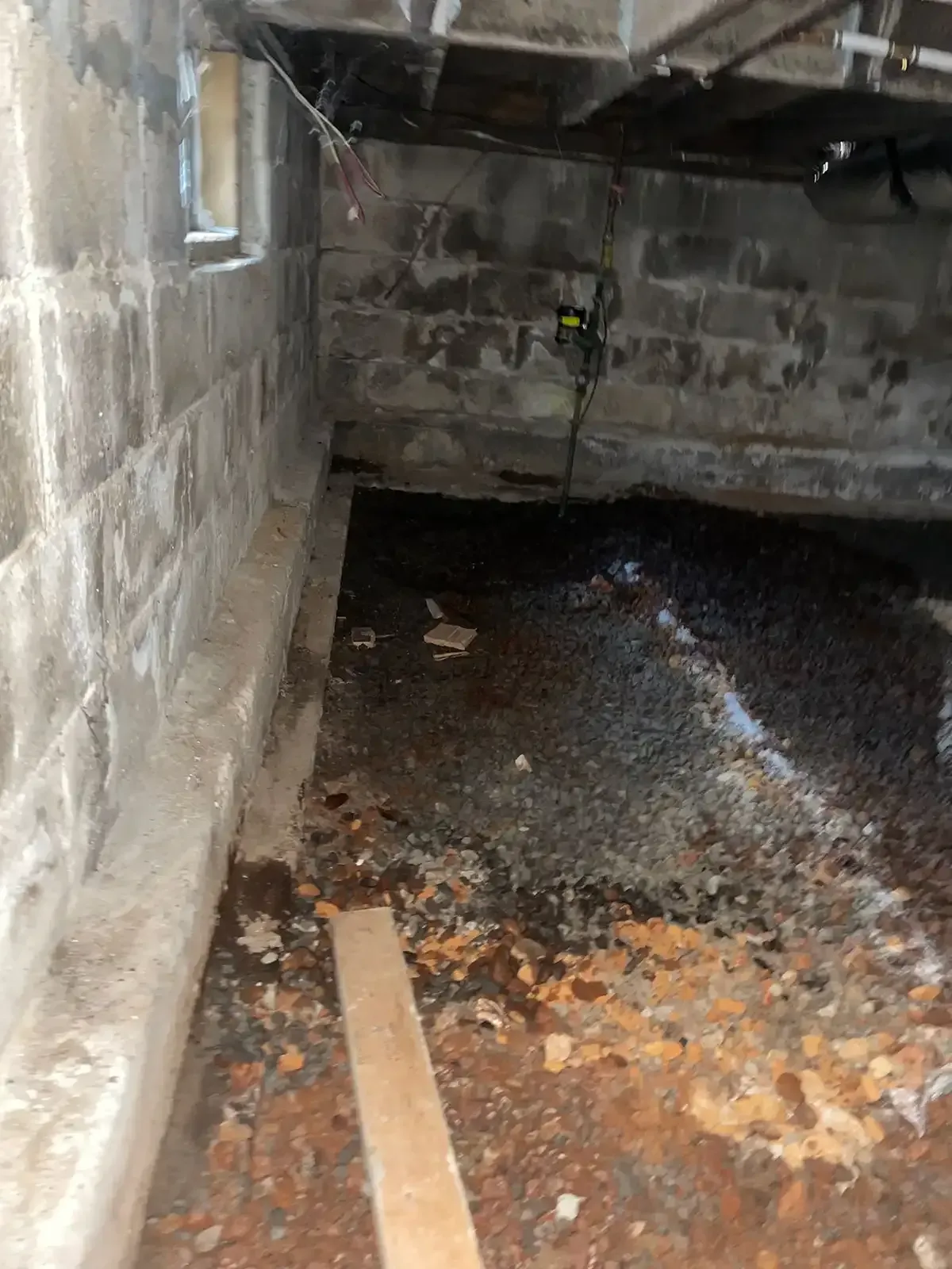 Damp concrete crawlspace with debris-covered floor, a small window, and exposed pipes.