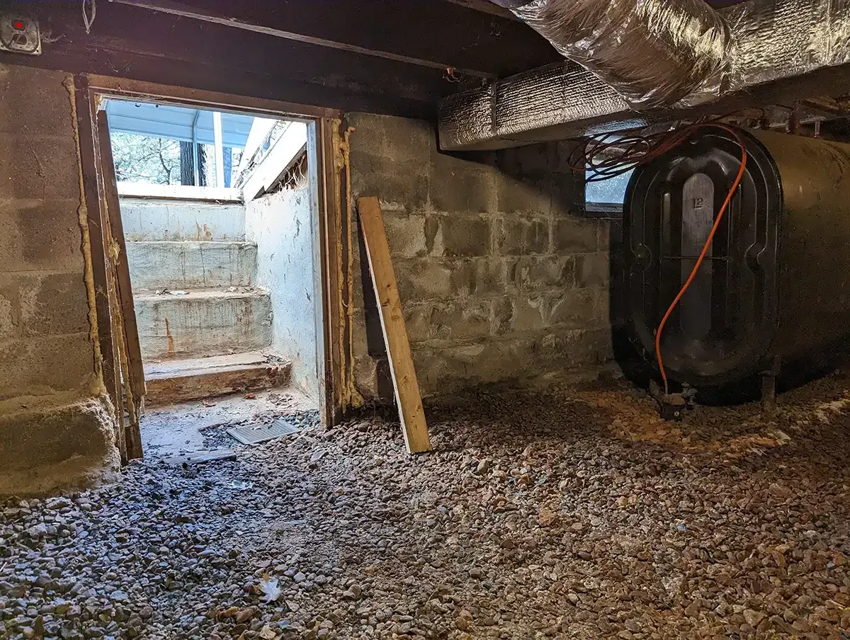Crawlspace with gravel floor, stone foundation, and a small doorway leading to concrete steps outside.