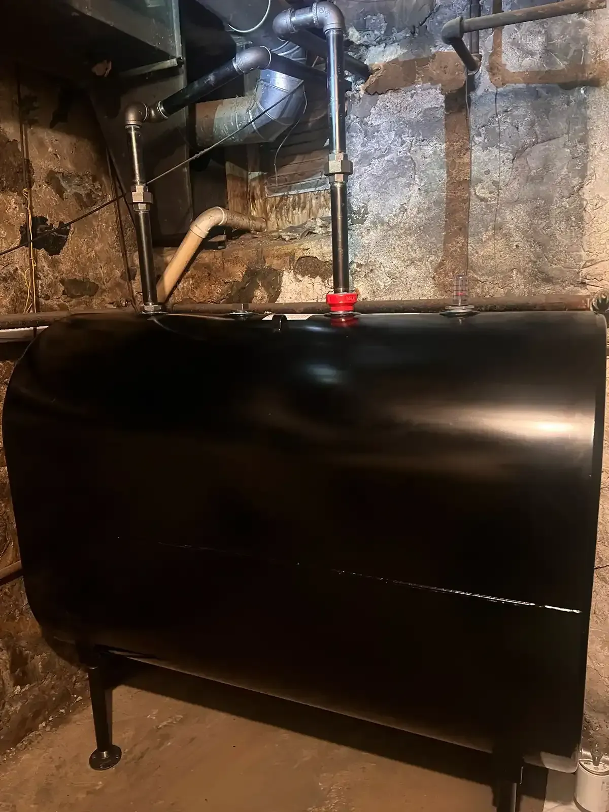 Black basement boiler or tank with pipes and valves against a stone wall.