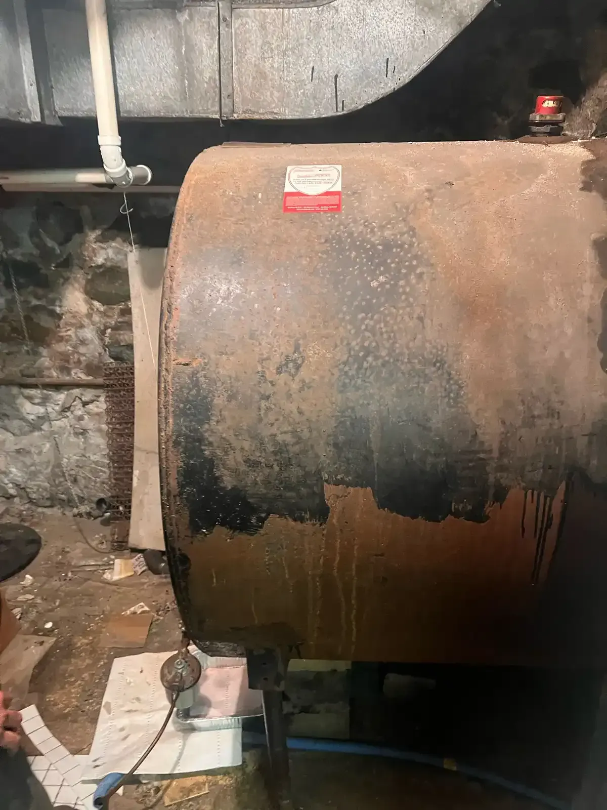 Rusty industrial tank in a dim basement, with a red warning label and pipes nearby.