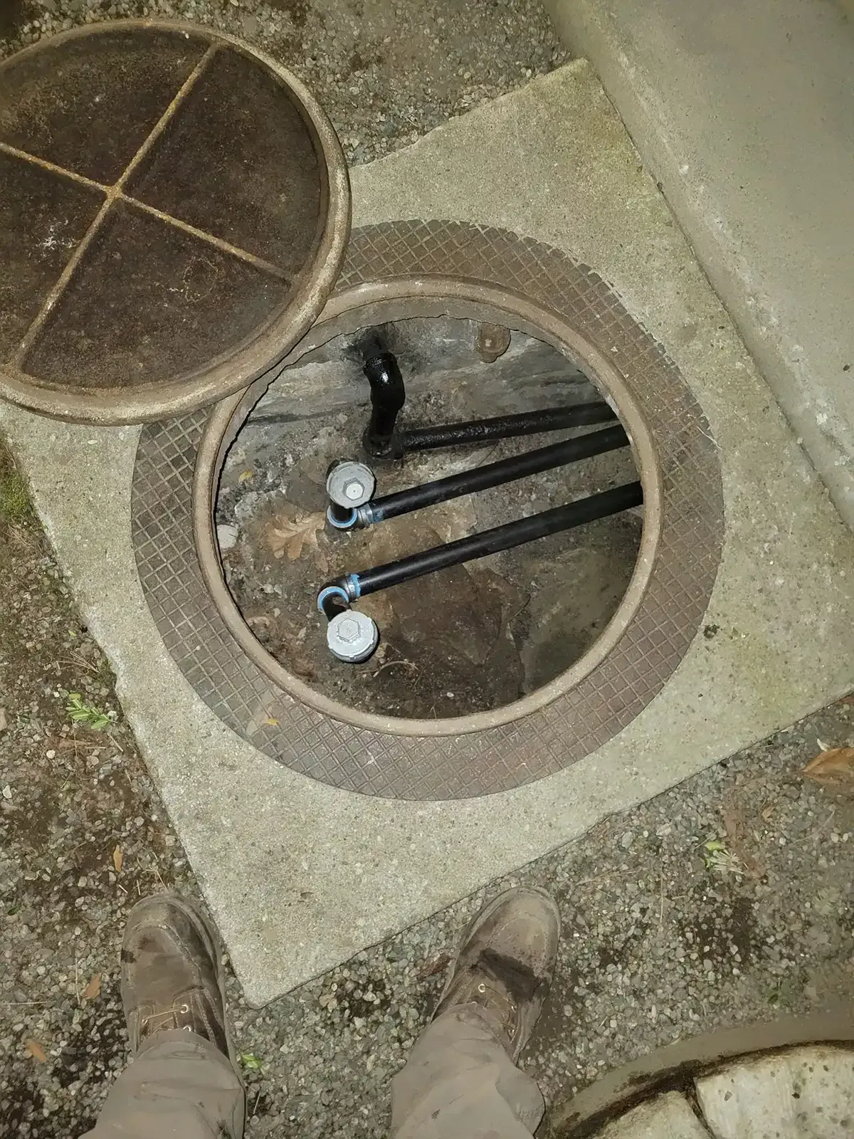 Open manhole with exposed pipes and valves in a concrete sidewalk, viewed from above.