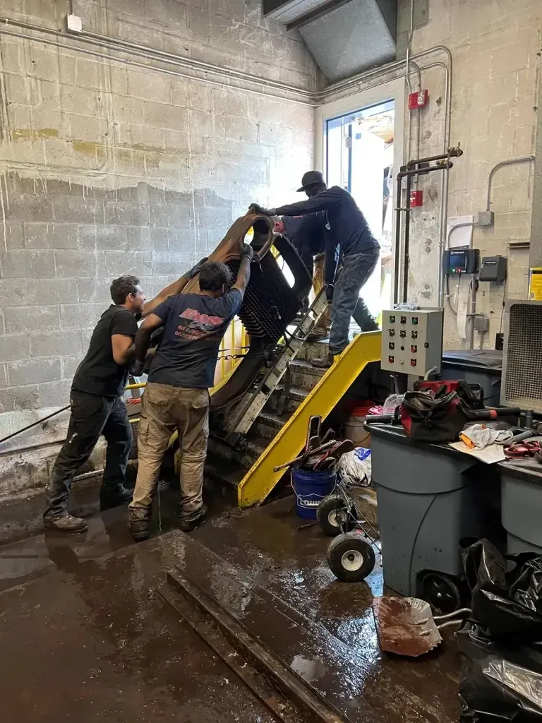 Three workers in a warehouse labor to maneuver a heavy piece of black machinery up a yellow-railed staircase.