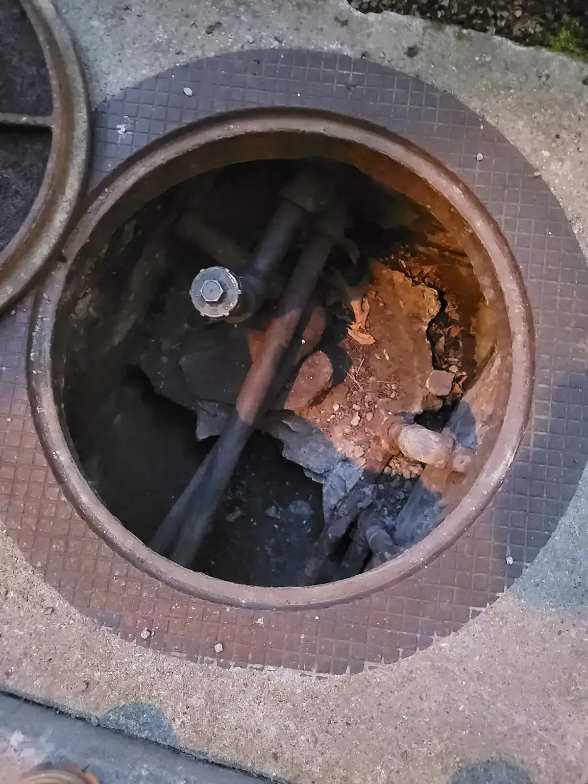 Open utility manhole with exposed cables and rust inside a concrete sidewalk.
