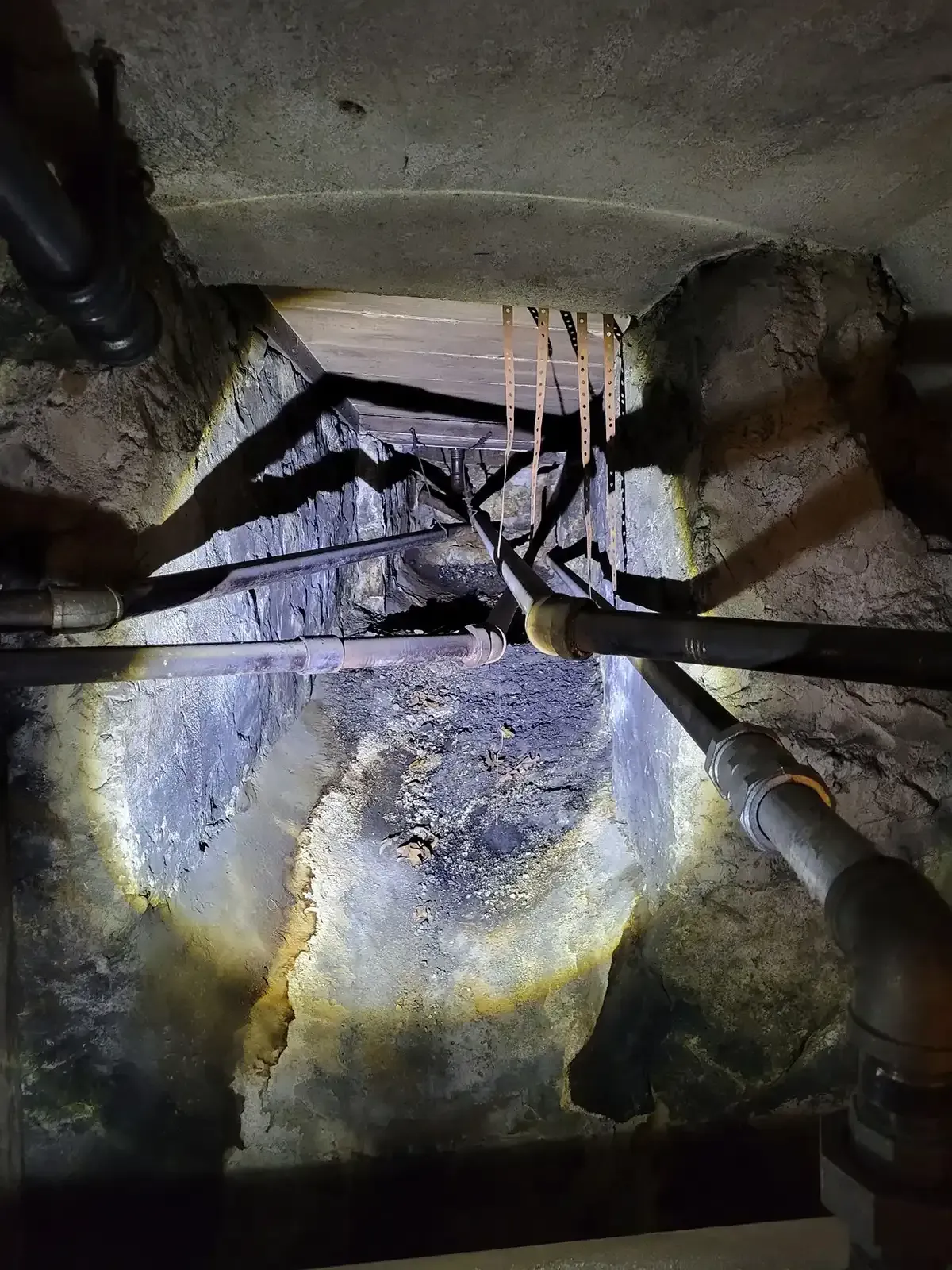 Dim industrial interior with pipes, wires, and a wet concrete wall lit by a flashlight.