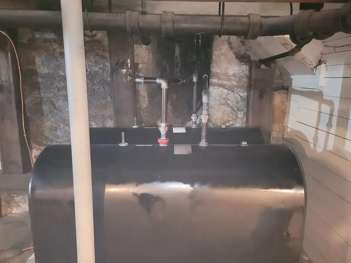 Black horizontal tank in a basement with connected pipes and valves against a stone wall.