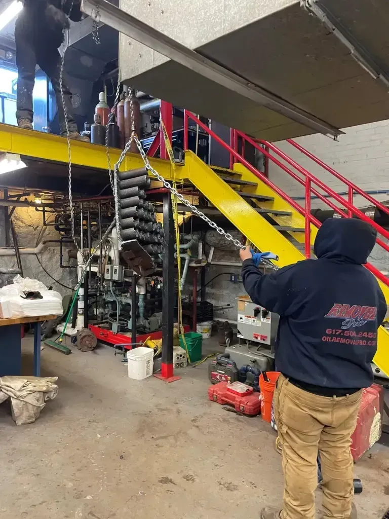 A worker in a dark hoodie uses a chain hoist to suspend a large metal object in a workshop with yellow stairs.