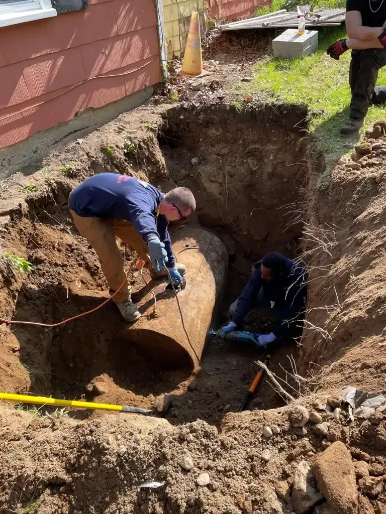 Two people work inside a deep trench, inspecting a large, rusted metal tank partially unearthed next to a house.