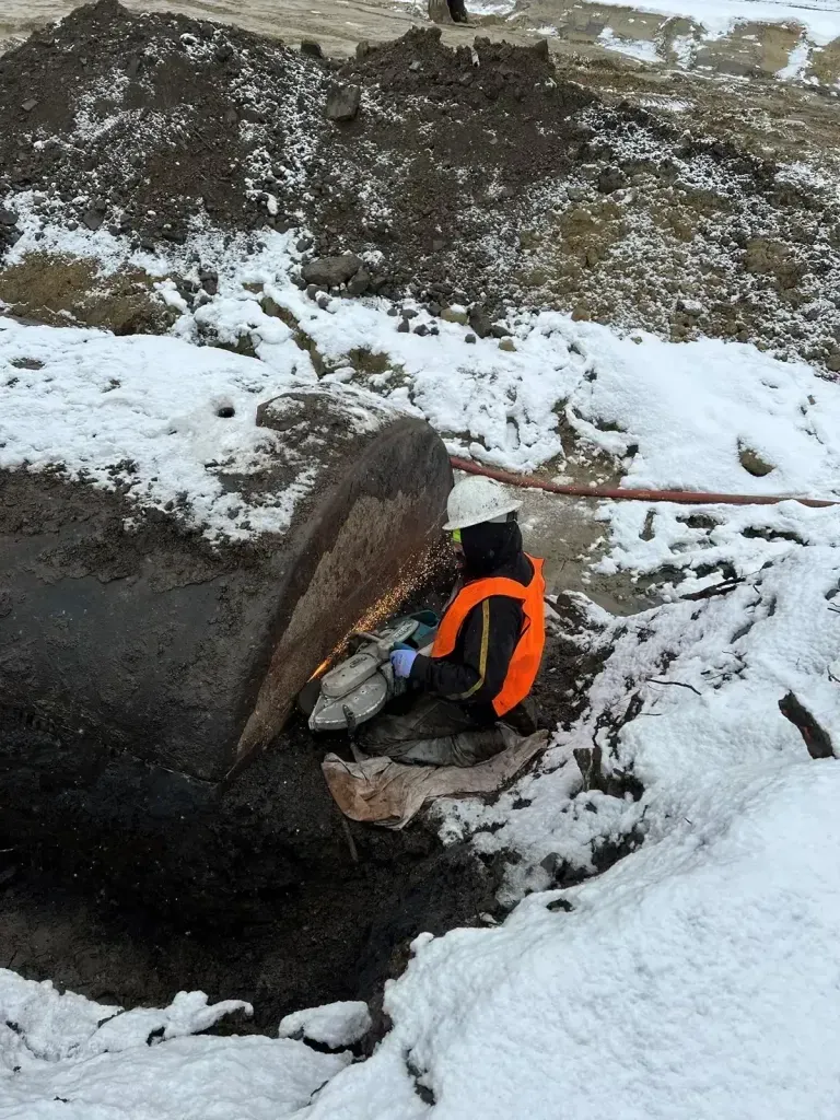A worker in a high-visibility orange vest uses a handheld power cutter on a large pipe in a snowy, outdoor trench.