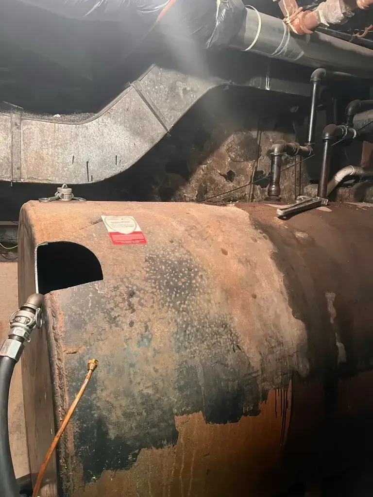 An old, rusty horizontal metal fuel oil tank with connected pipes in a dim basement setting.