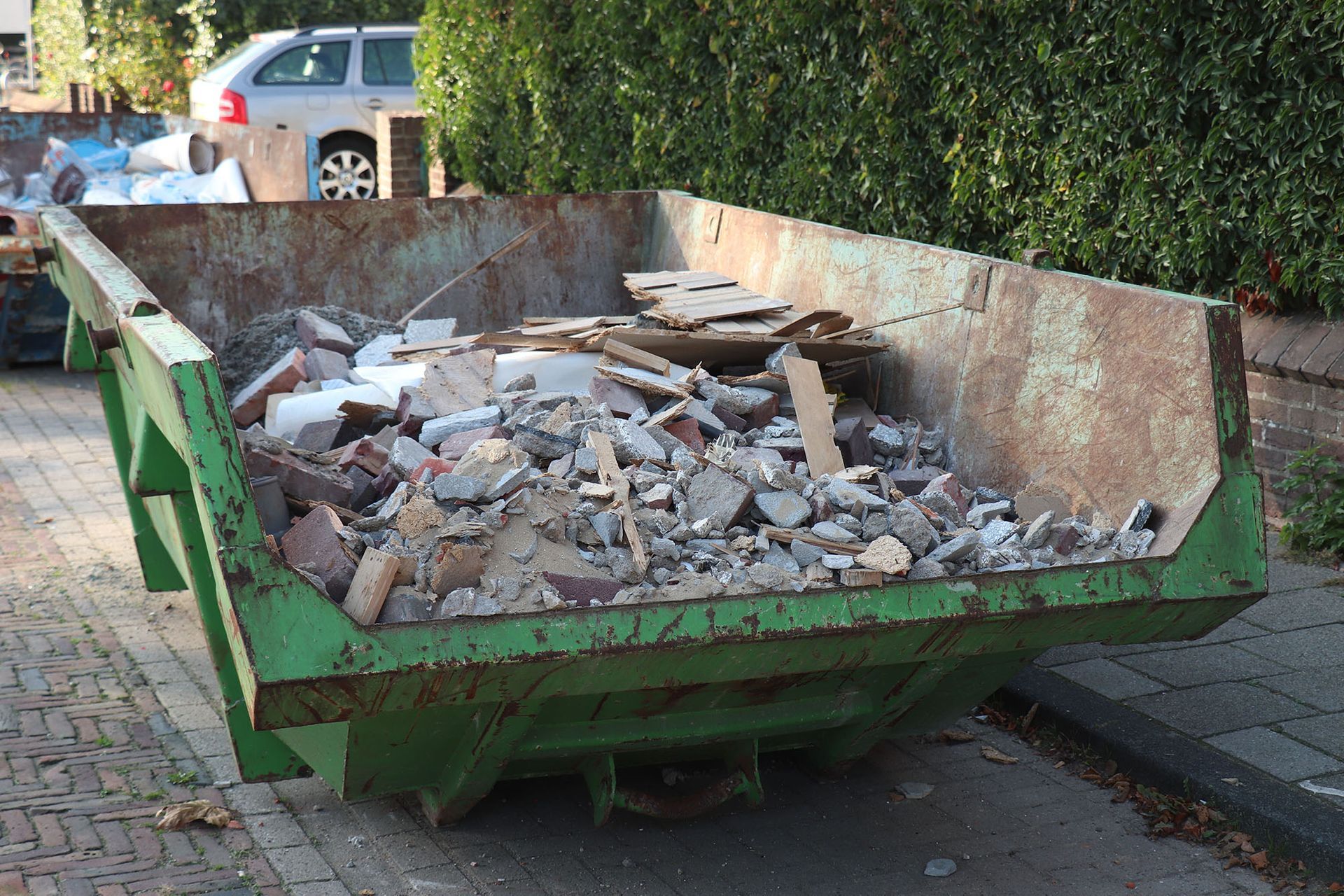 A green, weathered metal construction dumpster filled with building debris and rubble sits on a paved street.