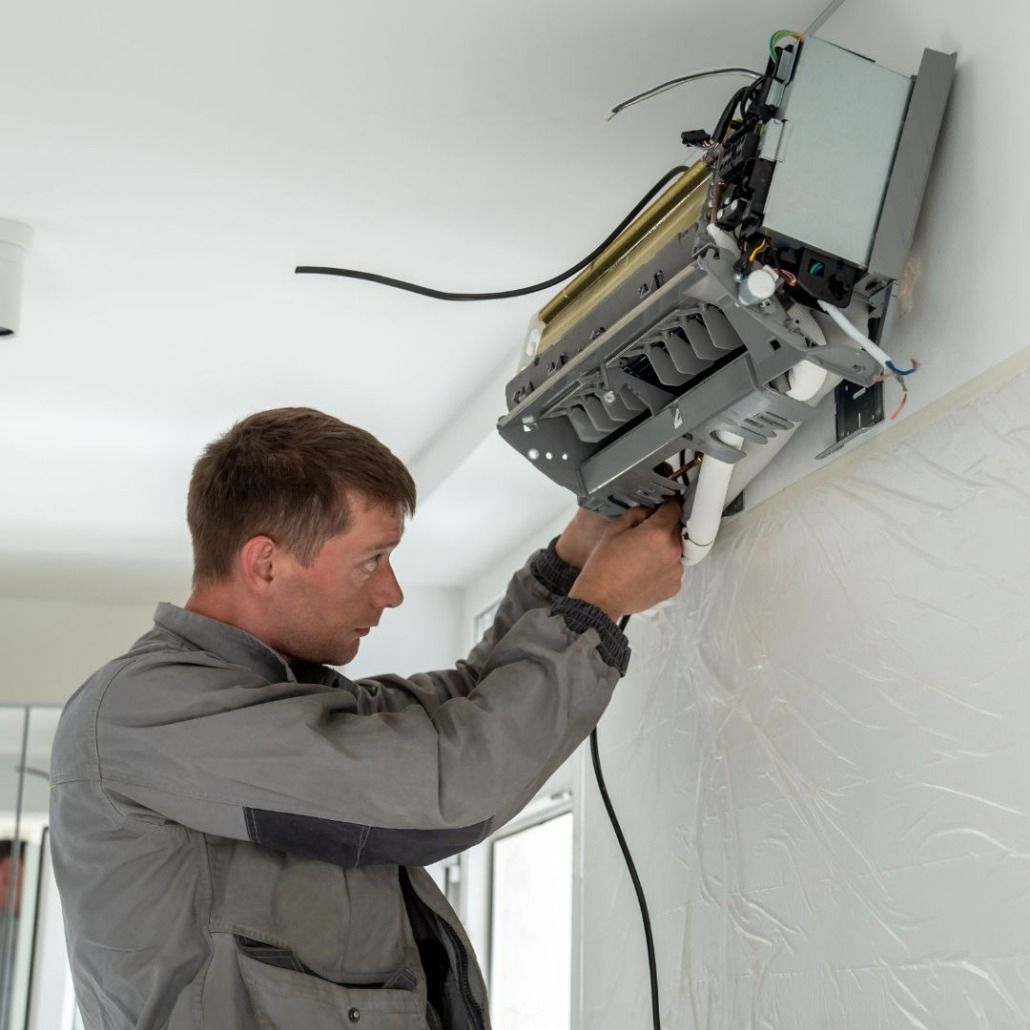A technician in a gray work jacket installs a wall-mounted air conditioning unit indoors.