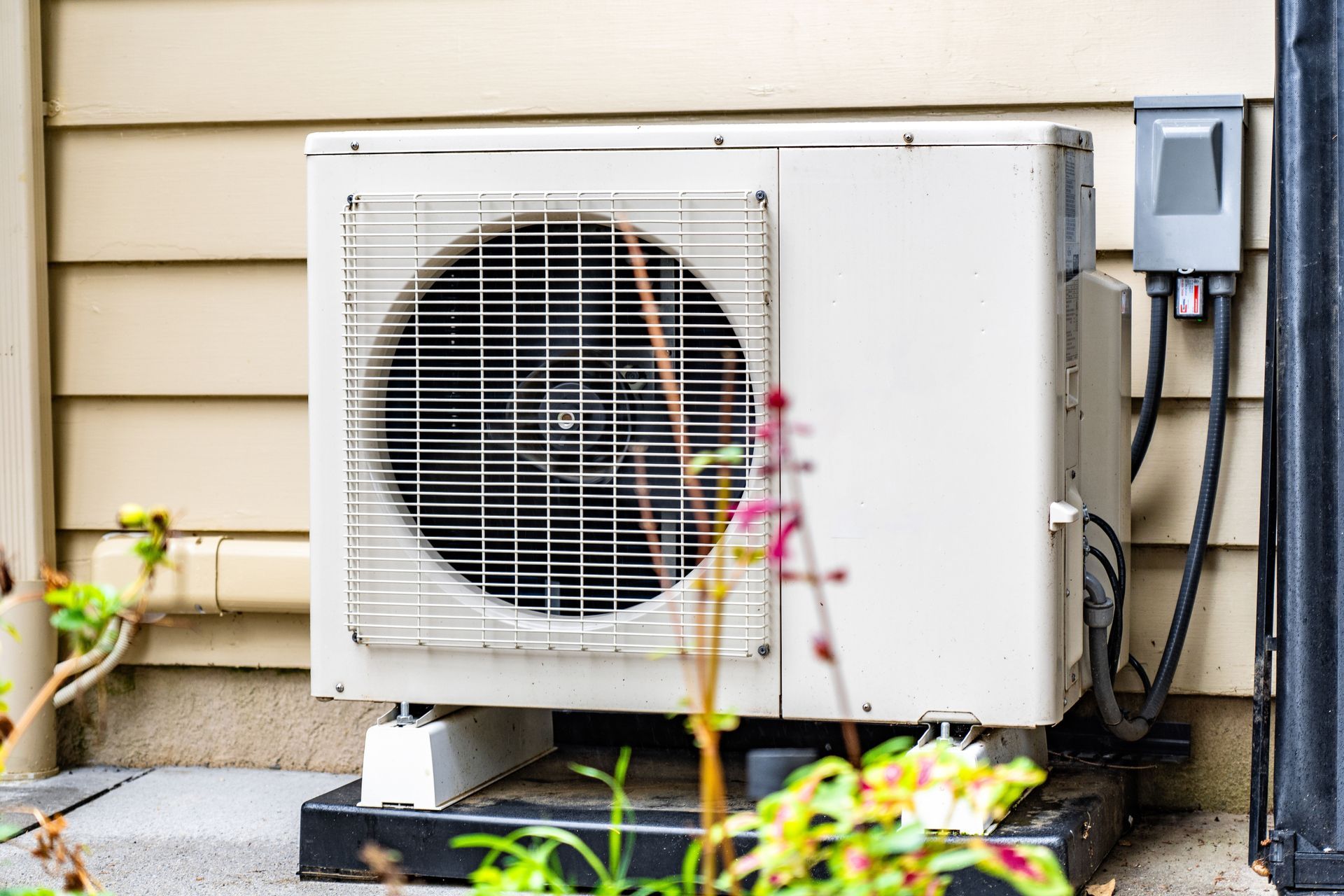 A tan outdoor air conditioning unit mounted on a black base against tan vinyl siding, with a gray electrical disconnect.