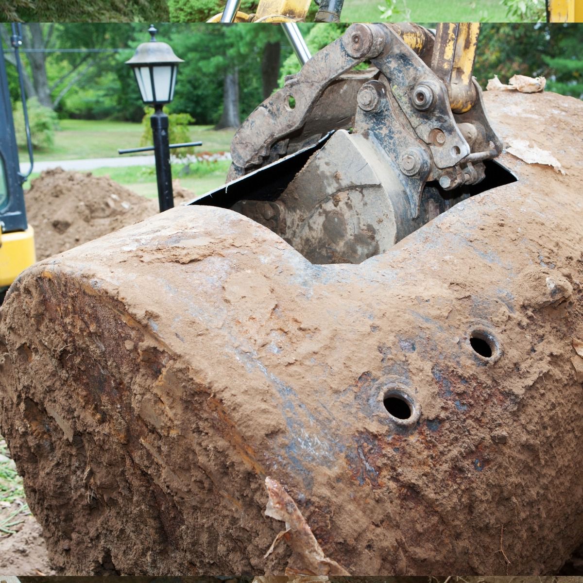 An excavator bucket cuts into a rusty underground storage tank in a grassy yard.