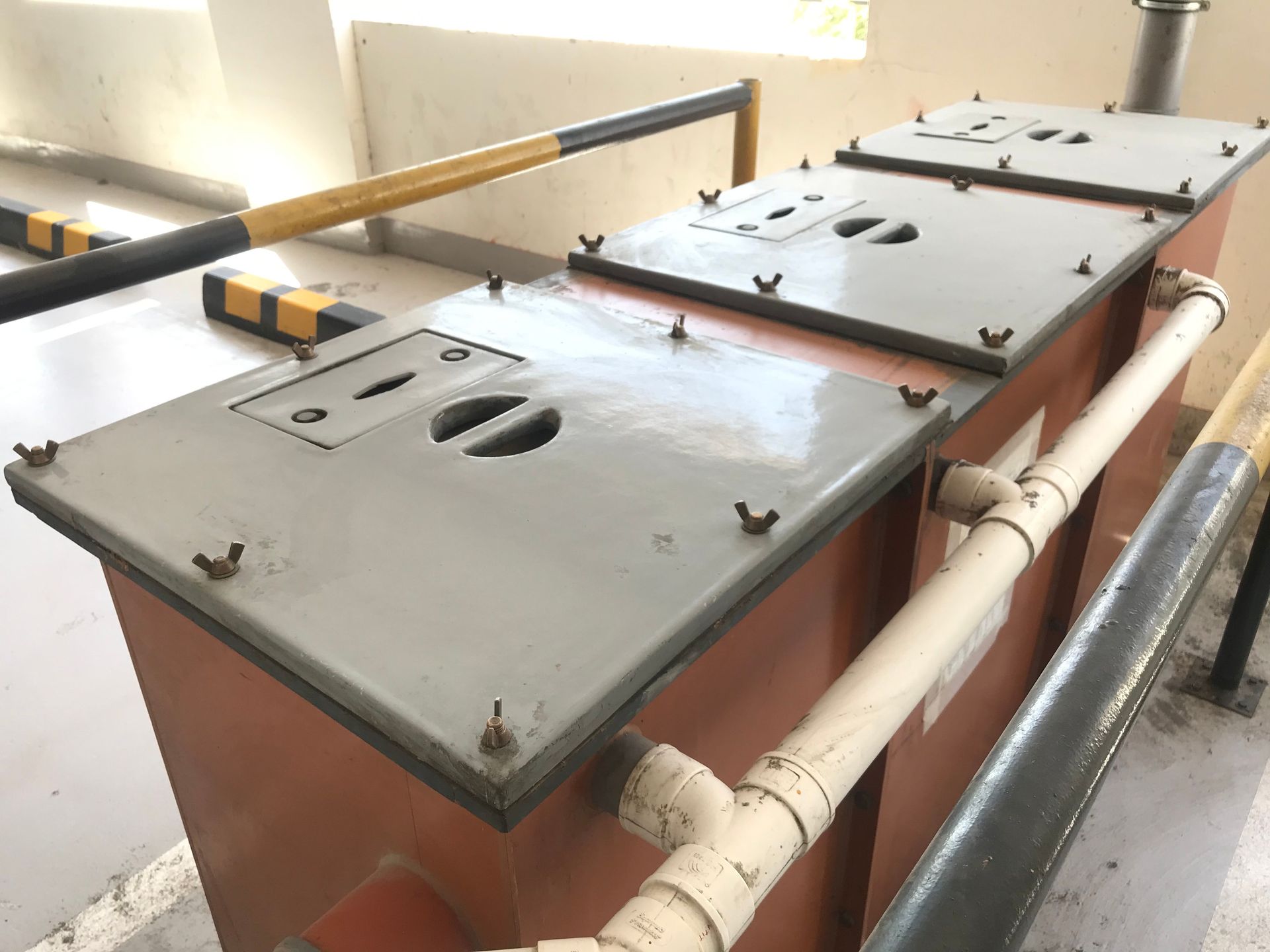 A row of three industrial grease traps with grey bolted lids and white PVC plumbing in a parking garage.