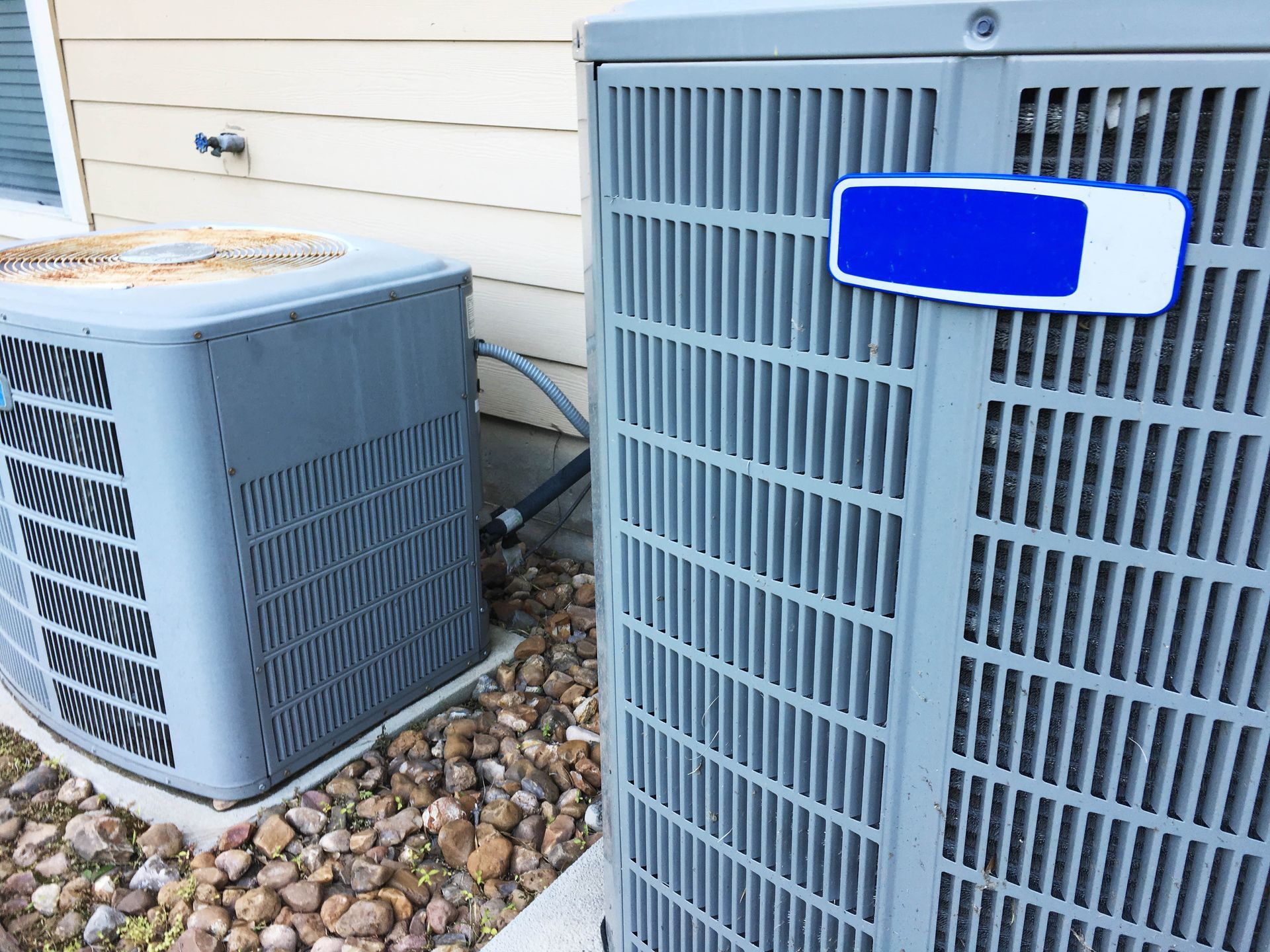 Two grey air conditioning units sit on a gravel bed next to the beige siding of a house.