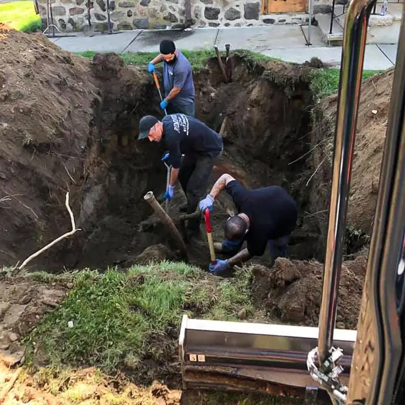 Three construction workers dig in a deep, dirt-filled pit near a stone building while an excavator bucket rests nearby.