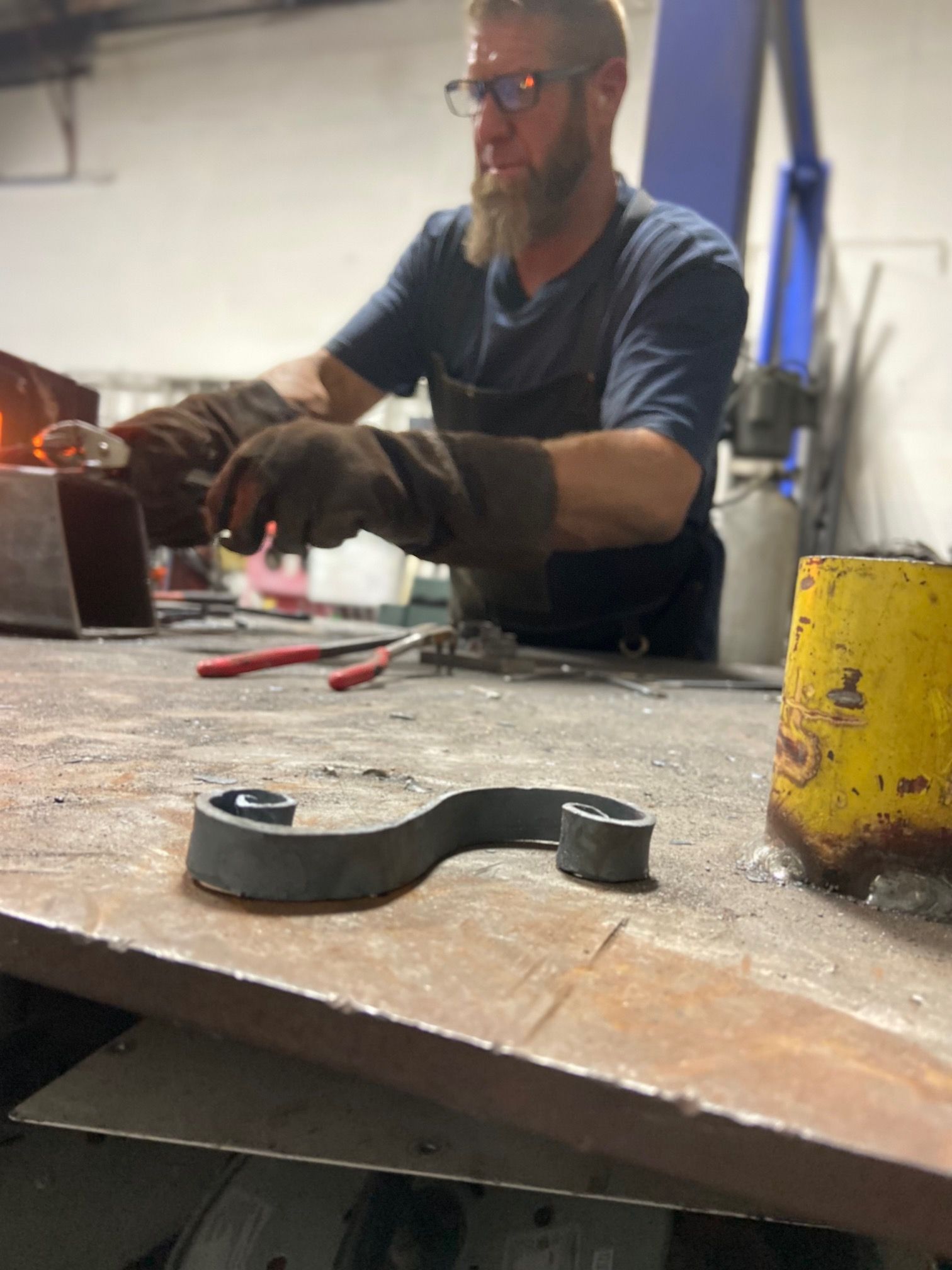 Blacksmith shaping metal. Man in shop, wearing gloves. S-shaped metal on table.