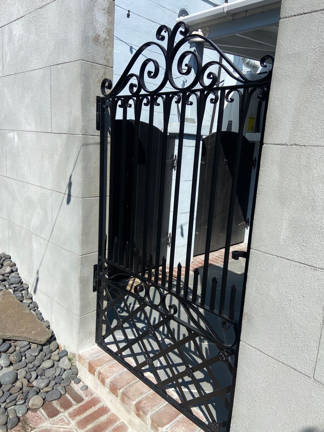 Black wrought-iron gate with ornate top, open against a brick and concrete wall.