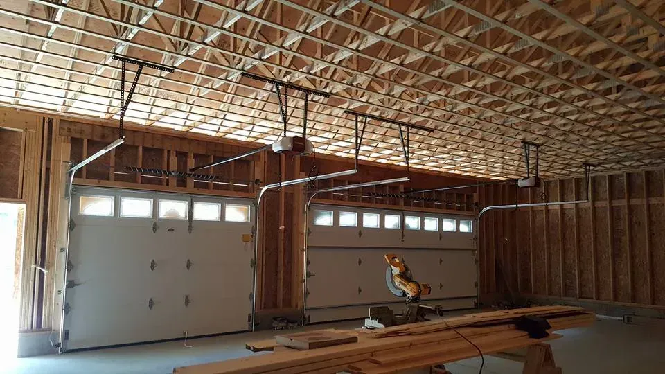 Interior view of a garage with two closed white doors, overhead garage door openers, and exposed wooden framing.