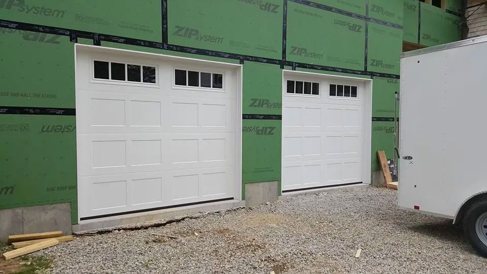 Two white garage doors with windows installed on a green-walled house under construction.