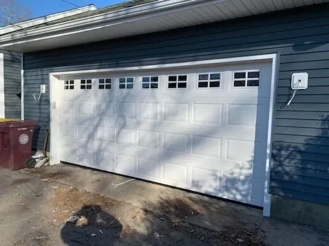 White garage door with small square windows above, beside blue siding, EV charger.