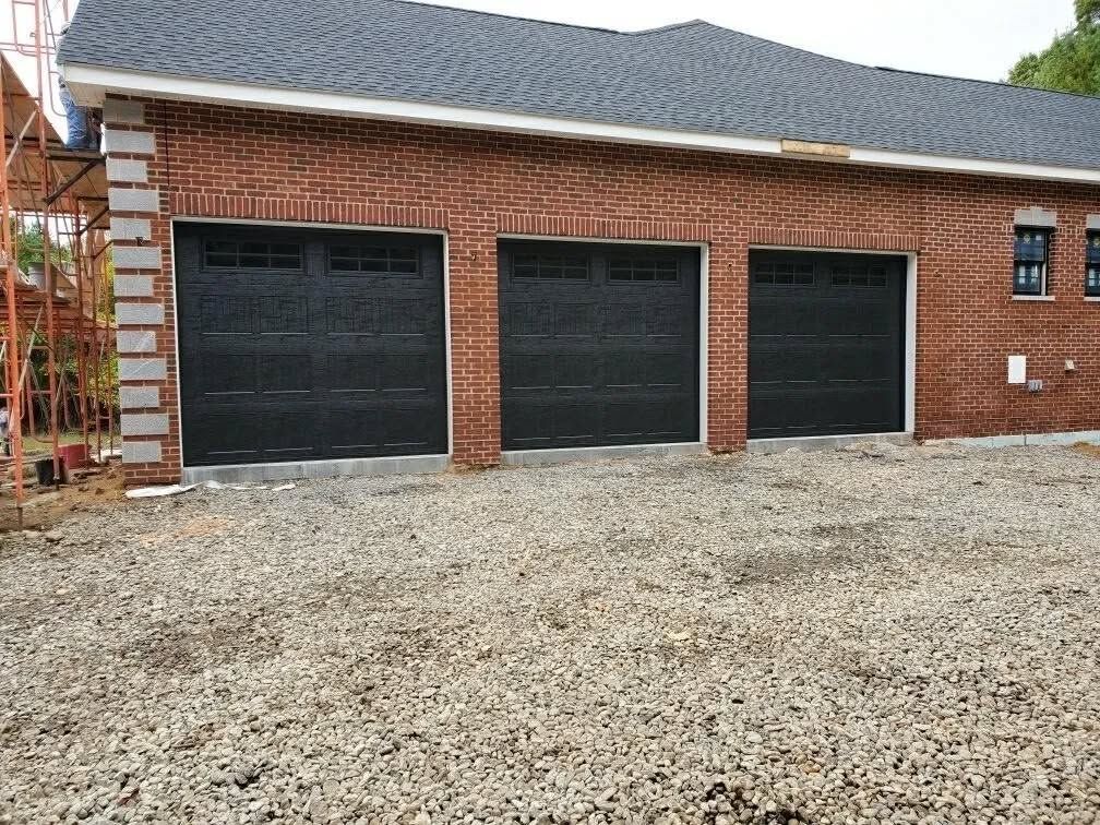 Three black garage doors on a brick building with a gravel driveway; construction scaffolding visible.