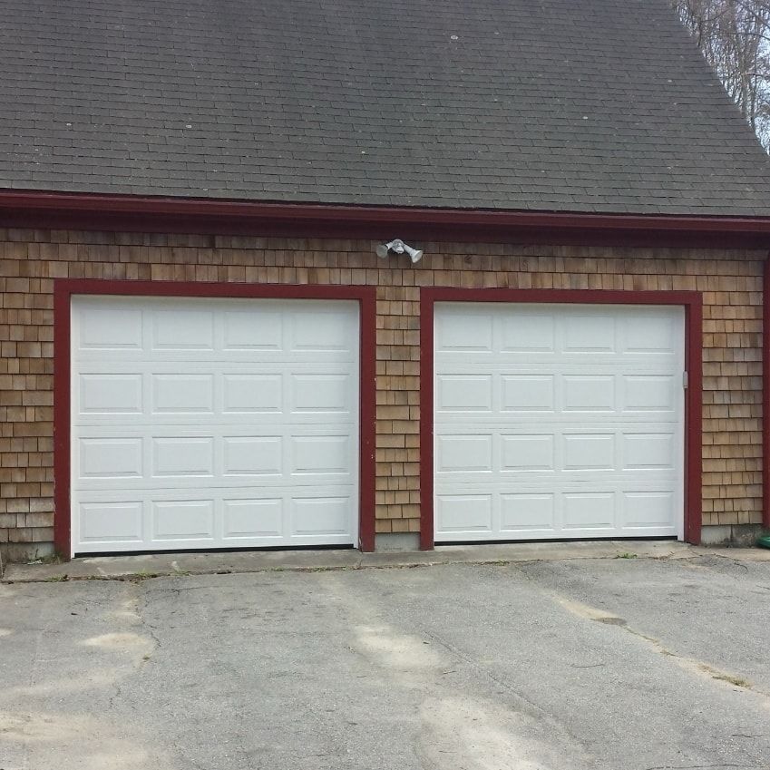 Two white garage doors with red trim, set in a brown shingled building with a gray driveway.