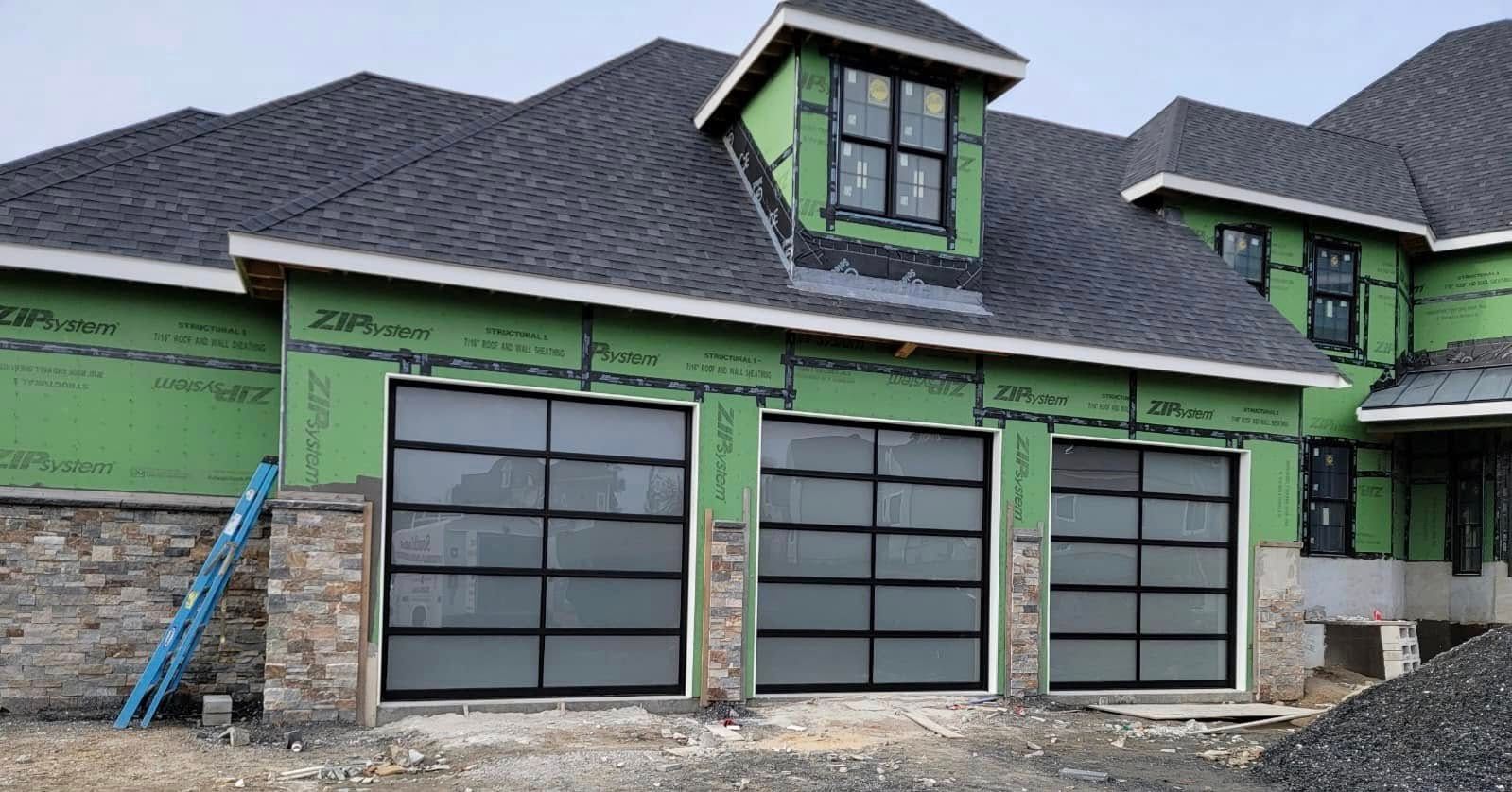 Three-car garage with glass doors, under construction. Stone pillars, green siding, and dark roof.
