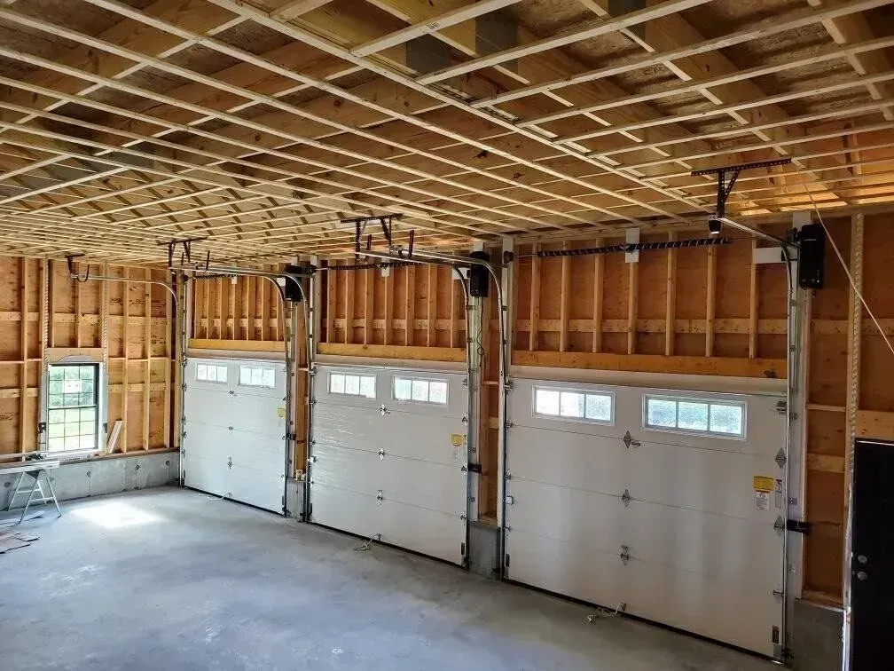 Interior view of a garage with three garage doors. Doors are white, framing is wood. Ceiling is unfinished.