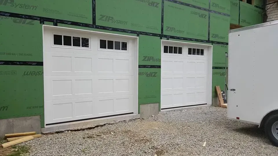 Two white garage doors with windows, against green siding, gravel ground, and a white trailer.