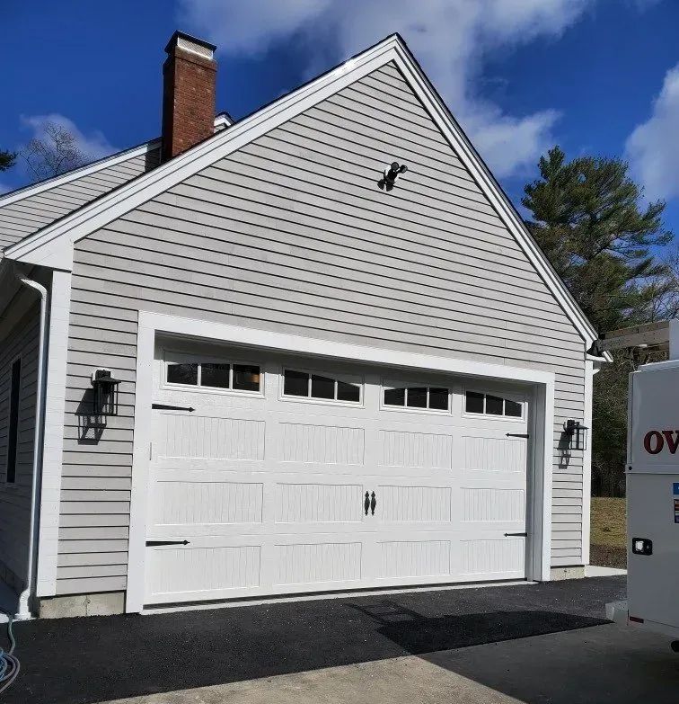 White garage door on a gray-sided house with black hardware and lights. Paved driveway.