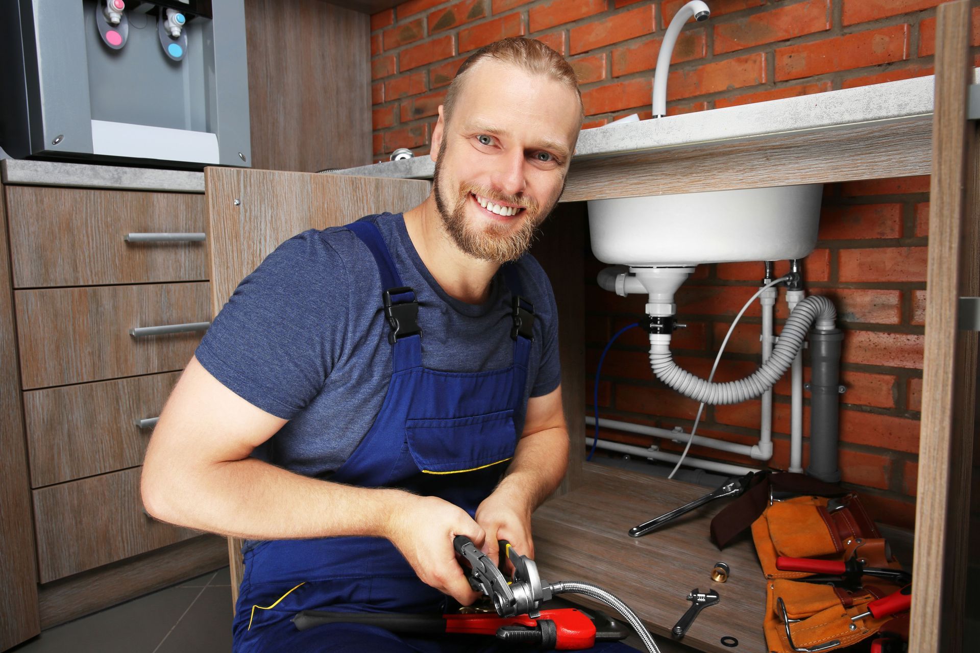 Plumber smiling, working under a kitchen sink, holding pliers.