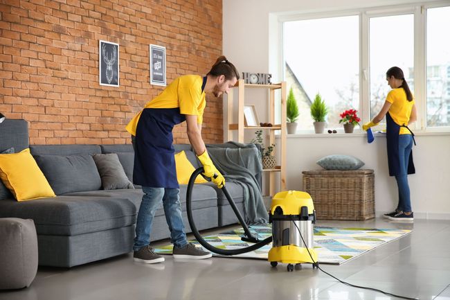 Two cleaners vacuuming a bright living room with a gray sofa and brick wall