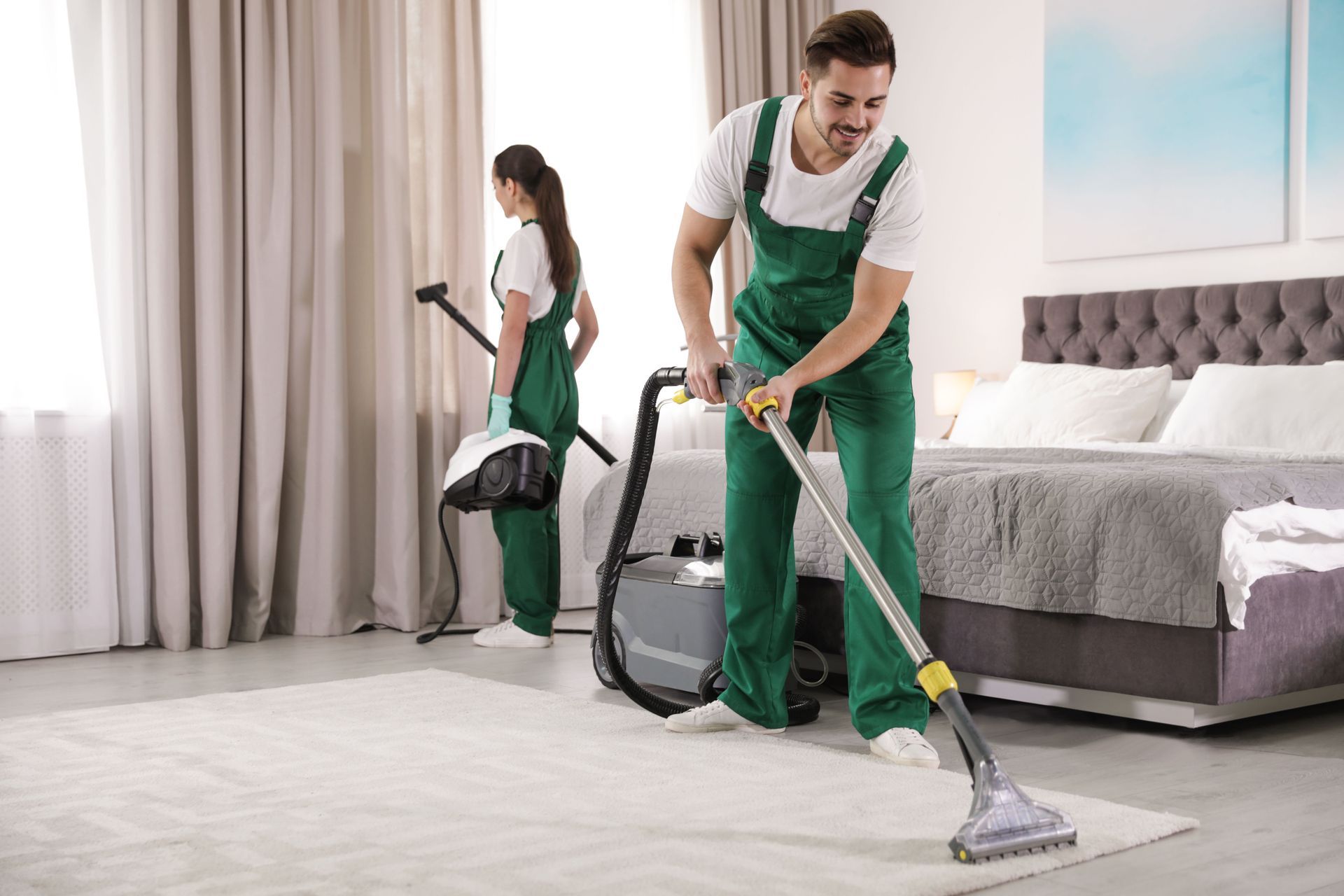 Two cleaners in green uniforms vacuuming a bright bedroom with a bed and curtains