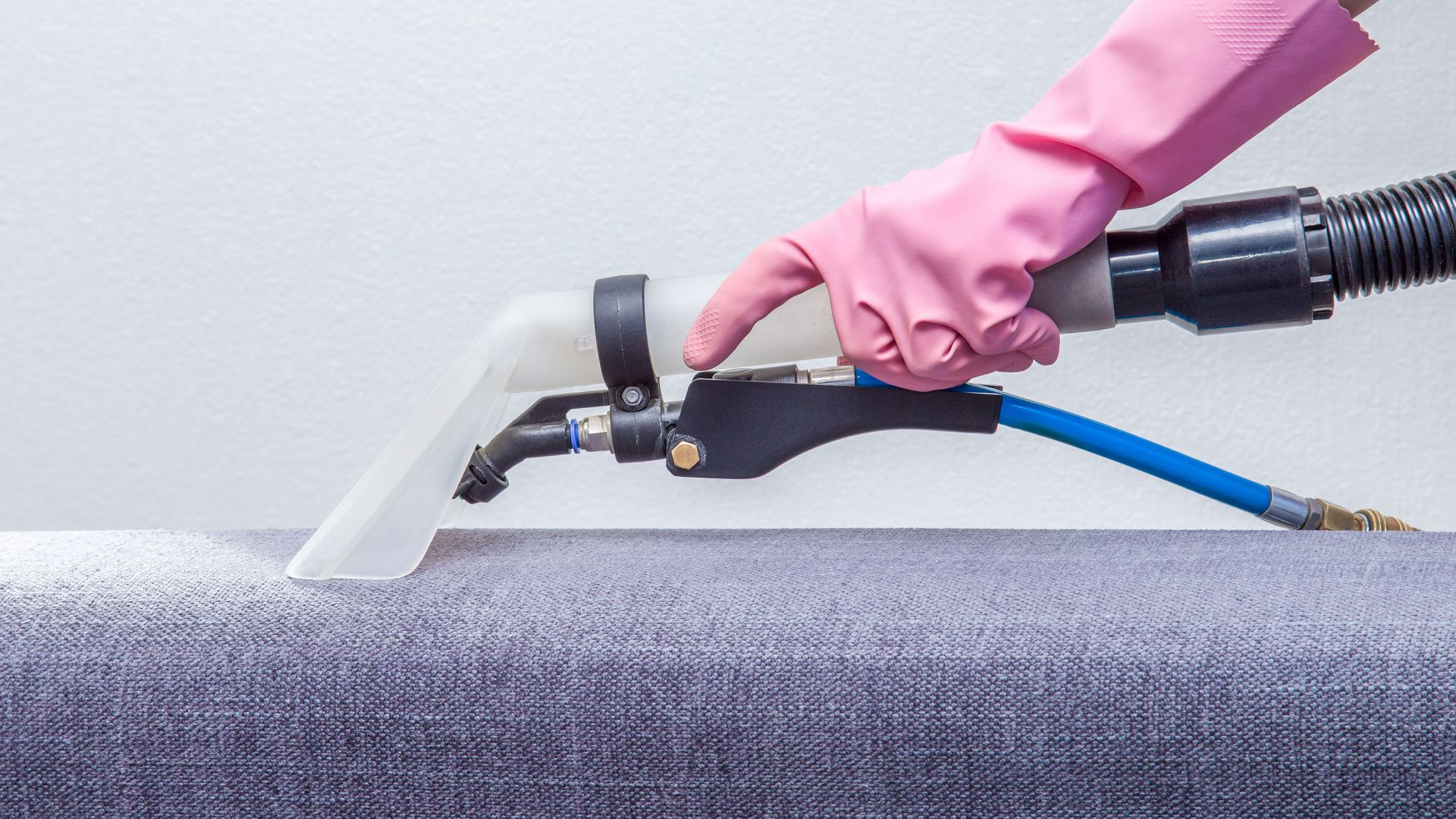 A pink-gloved hand uses a vacuum tool to clean a gray upholstered couch.