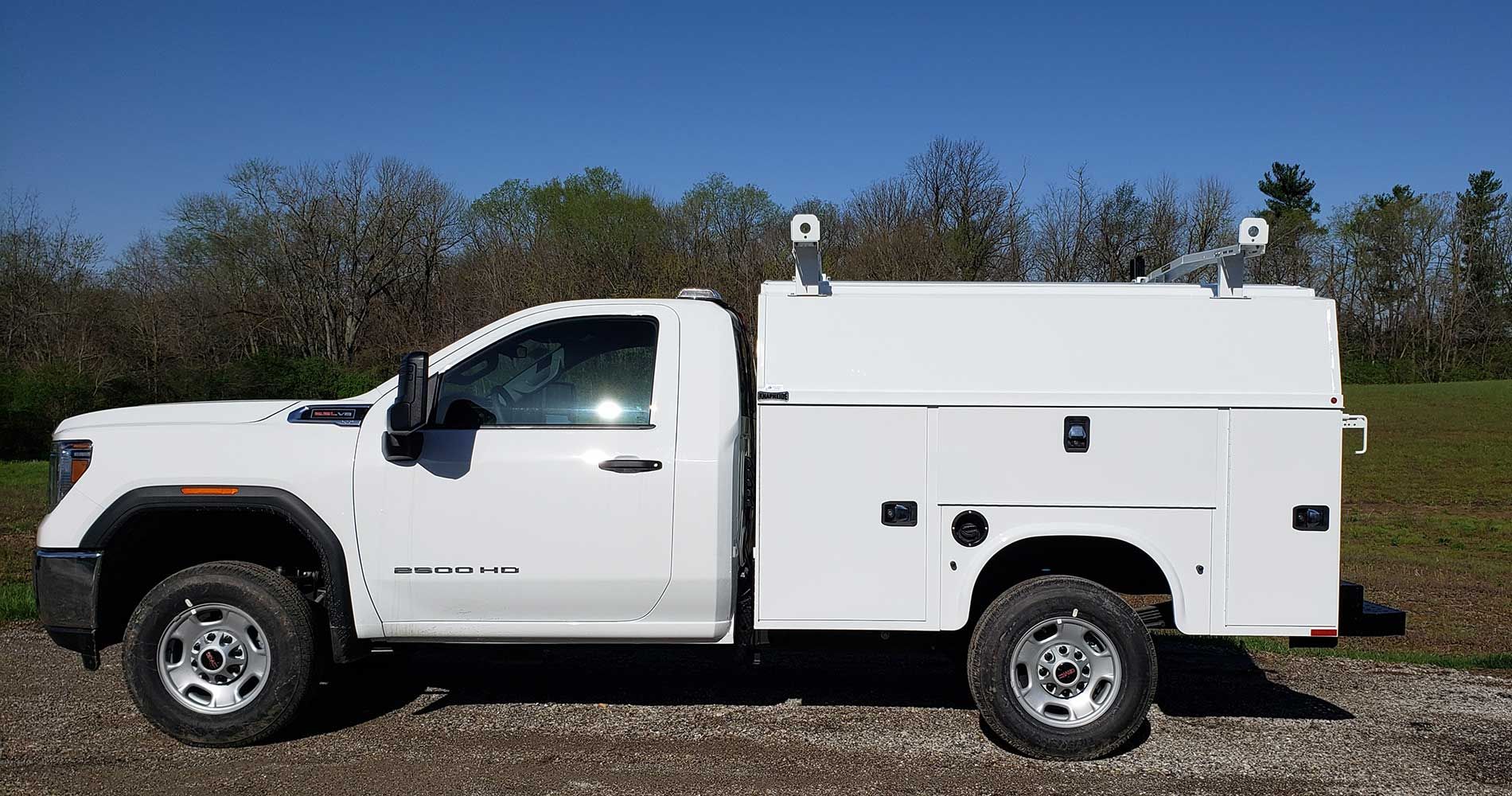 White work truck with enclosed bed, on a gravel drive with a green field and trees in the background.