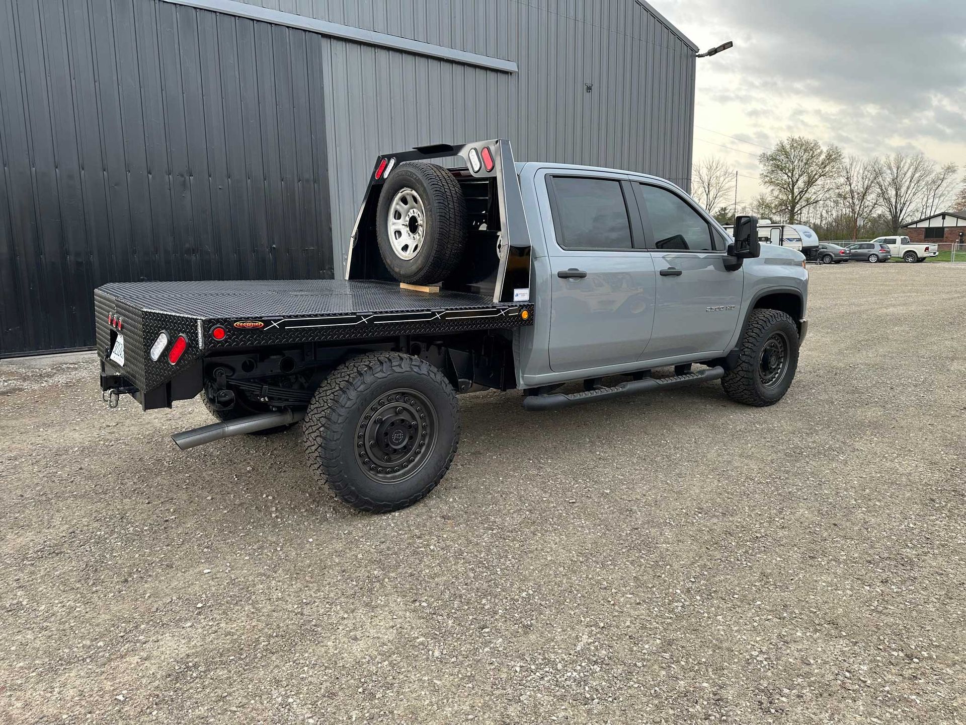 Silver flatbed pickup truck on gravel, spare tire mounted on cab guard.